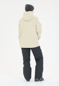 Wasserfeste beiger Jacke mit Kapuze, kombiniert mit schwarzen Hosen und schwarzen Stiefeln. Glatte Textur, minimalistische Gestaltung und funktionales Material.