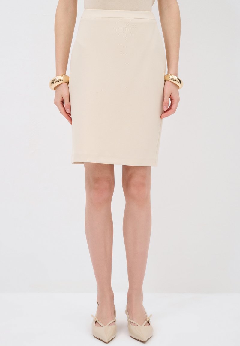 Falda de lápiz beige con un patrón texturizado, corte recto y cintura de altura media. Combinada con tacones nude de punta y una pulsera dorada en la muñeca.