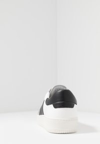 Vit sneaker med svarta läderdetaljer, texturerad sula, rundad form och vadderad krage. Har varumärkeslogotyp på hälen.