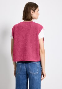 Fleeceväst i starkt rosa med en texturerad yta, med lös passform och utan ärmar, bärs över en vit skjorta och matchas med blå jeans.