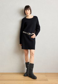 Vestido de punto negro de gran tamaño con escote redondo, combinado con un cinturón negro con tachuelas y botas de cuero negras hasta la rodilla. Textura suave, diseño moderno.