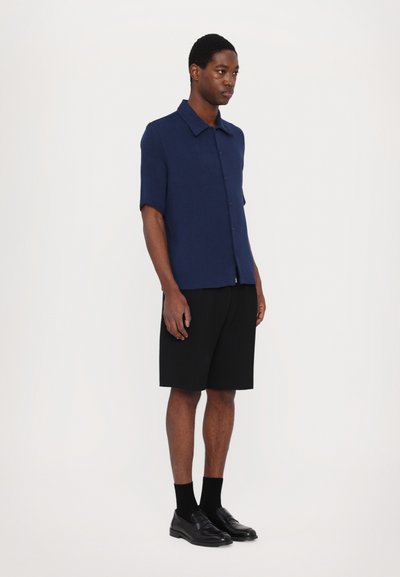 Short-sleeve navy blue button-up shirt made of lightweight fabric, worn with black shorts and black slip-on shoes. Simple, modern design.