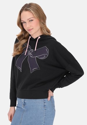 Felpa nera con un grande contorno bianco di un disegno a fiocco, cordini rosa e una vestibilità rilassata, abbinata a una gonna di jeans.
