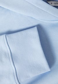 Svijetloplavi pulover s glatkom teksturom i rebrastim rukavima. Ima v-izrez i savijeni ovratnik za opuštenu fit.