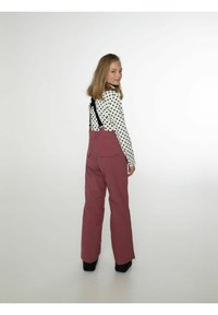 Bordo lumppuhousut olkahihnoilla, joiden istuvuus on löysä, sisältäen kaksi takataskua ja mustia yksityiskohtia. Päällä pitkähihaisesta pilkullisesta paidasta.