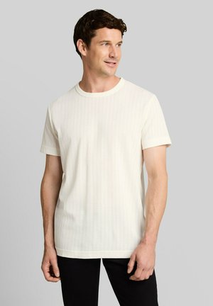 Man draagt een effen wit T-shirt met korte mouwen en een zwarte broek, kijkt opzij tegen een grijze achtergrond.