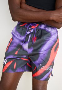 Badräktsbyxor i ett lätt tyg med ett djärvt abstrakt mönster i lila, rött och svart; inkluderar dragsko i midjan och logotypdetalj.