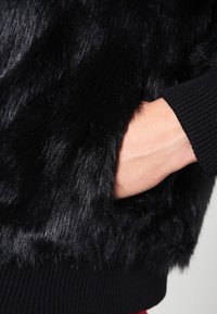 Svart fuskpälsjacka med en mjuk, texturerad yta och ribbade svarta muddärmar. En hand visas vila i en sidoficka.