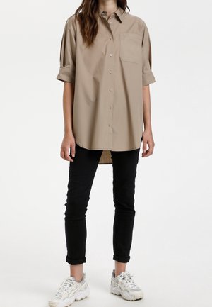 Beżowa koszula oversize z krótkimi rękawami, zapinana na guziki z przodu, z jedną kieszenią na piersi i luźnym fasonem, w zestawie z czarnymi, podwiniętymi spodniami oraz trampkami.