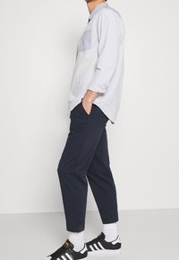 Ljust grå långärmad skjorta med knappstängning och bröstficka, kombinerad med marinblå byxor och svarta sneakers med vita detaljer.