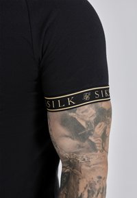 Sort kortærmet skjorte med en tætsiddende pasform, der har en fremtrædende manchet i sort og guld med logo. Stoffet ser blødt ud og har en let glans.