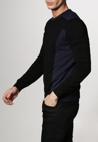 Pull à motifs noirs et bleu marine, col rond, avec des panneaux de couleur contrastante sur les côtés et des poignets côtelés. Matière douce avec une coupe ajustée.