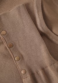 Tissu tricoté marron clair avec un poignet côtelé présentant quatre boutons ronds mats le long de la manche.