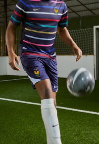 Fotbalový dres Nike se vyznačuje vodorovnými pruhy v námořnické modré, růžové, žluté a modré barvě. Kraťasy mají logo Coq a bílé akcenty. Hráč kopne šedý míč.