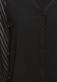 Zwarte blouse met een v-hals, knoopsluiting en een getextureerde voorkant, met mouwen in een geribd patroon voor contrast en detail.