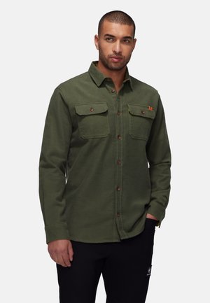 Green flannel shirt with two chest pockets, brown buttons, and a collar. Soft texture, casual design, worn over black pants.
