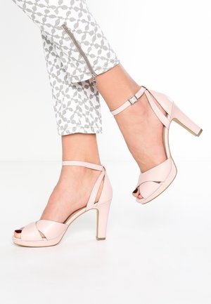 Feet wearing pale pink high-heeled sandals with ankle straps paired with white pants featuring gray leaf patterns and a zipper detail.