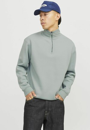Light green fleece pullover with a high collar and half-zip. Features ribbed cuffs, contrasting seams, and worn with dark denim pants.