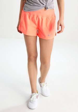 Personne portant un short de sport corail, un haut gris noué et des chaussures de course blanches, debout sur un sol blanc.