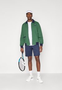 Casaco de malha verde com colarinho azul-marinho, t-shirt branca, calções azul-marinho e ténis brancos. A segurar uma raquete de ténis com detalhes azuis.