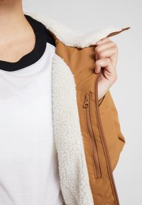 Brązowa kurtka ocieplana z białym futerkiem, z boczną kieszenią na zamek i czarnym kołnierzykiem. Nosiona na białej koszuli.