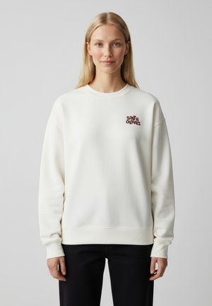 Vrouw met lang blond haar, gekleed in een witte sweater met de tekst "Salt & Olives" op de borst en een zwarte broek, staand tegen een effen achtergrond.
