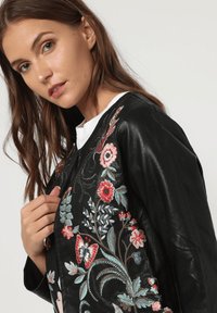 Chaqueta de cuero negro con bordados florales coloridos en rojo y azul. Diseño con cuello redondo y ajuste holgado.