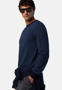 Marinebluse mit Streifen und langen Ärmeln, Rundhals-Ausschnitt, kombiniert mit dunklen Shorts und einem um die Taille gebundenen Sweatshirt. Model trägt Sonnenbrille.
