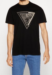 Schwarzes Baumwoll-T-Shirt mit einem silbernen dreieckigen Logo, das "GUESS" und ein Fragezeichen zeigt, rundem Ausschnitt und kurzen Ärmeln.