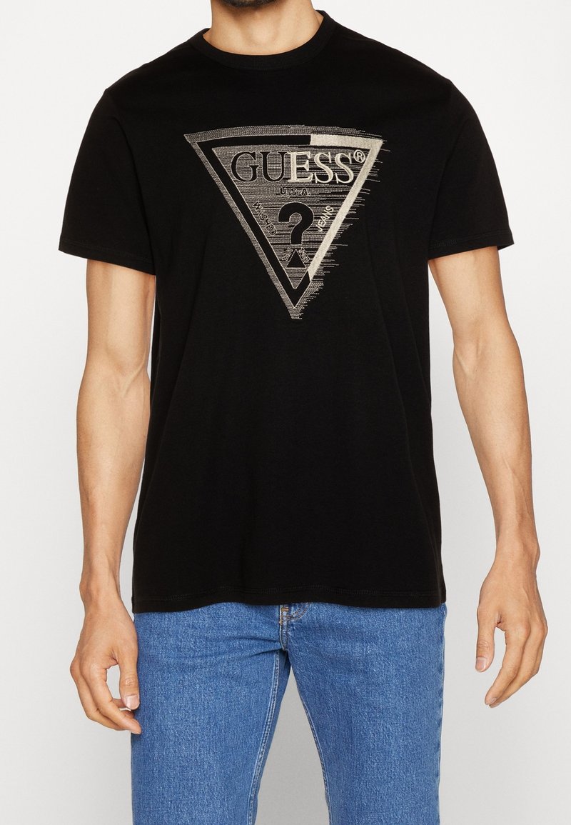 Schwarzes Baumwoll-T-Shirt mit einem silbernen dreieckigen Logo, das "GUESS" und ein Fragezeichen zeigt, rundem Ausschnitt und kurzen Ärmeln.