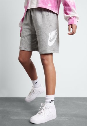 Person trägt graue Nike-Shorts, weiße Nike-Sneakers, weiße Nike-Socken und ein rosa Batik-Langarmshirt und steht auf grauem Boden.