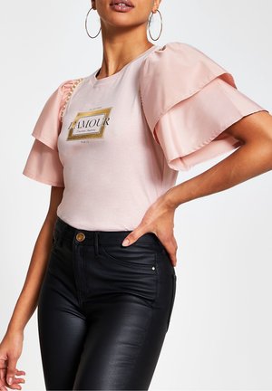 Vrouw draagt een lichtroze top met grote gelaagde ruches aan de mouwen en de tekst "L'AMOUR", gecombineerd met een zwarte broek met hoge taille.