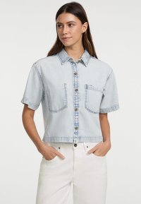 Hellblaue, kurz geschnittene Denim-Jacke mit kurzen Ärmeln, Holzknöpfen, zwei Brusttaschen und kontrastierendem Stitching. Glatte Textur.