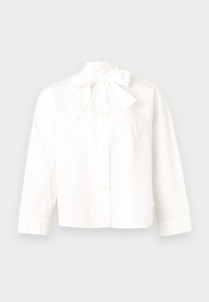 Blouse blanche à manches longues avec boutons, ornée d'un grand nœud noué au col, présentée sur un fond neutre et à plat.
