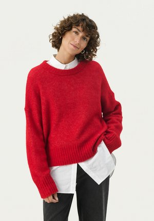 Rode gebreide trui met een brede ronde halslijn, geribbelde onderrand en manchetten, gedragen over een witte blouse met kraag, gecombineerd met zwarte broeken.