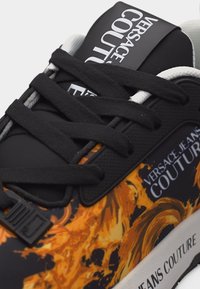 Μαύρο αθλητικό παπούτσι με πορτοκαλί φλόγες, μαύρα κορδόνια και λευκή επωνυμία "Versace Jeans Couture" στη γλώσσα και στο πλάι.