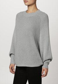 Světle šedý oversized pletený svetr s žebrovanou strukturou, kulatým výstřihem a dlouhými rukávy, v kombinaci s černými přiléhavými kalhotami.