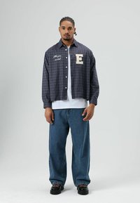 Overgrote geruite blouse met knopen in blauw, rood en wit, met een geborduurde "E" en tekst. Gecombineerd met loszittende blauwe spijkerbroek en zwarte schoenen.