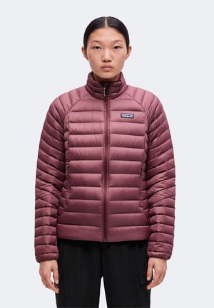 Žena s tmavými vlasmi oblečená v mauve prešívanej bunde Patagonia so zipsom vpredu, stojaca na jednoduchom svetlom pozadí.