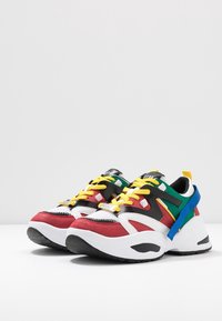 Zapatillas coloridas que presentan una mezcla de materiales rojos, negros, blancos y verdes, con cordones amarillos y una suela texturizada. El diseño incluye acentos y capas.