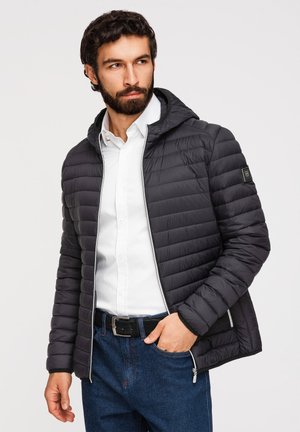 Mann mit Bart trägt schwarze gesteppte Jacke über weißem Hemd und blauen Jeans, schaut seitlich mit einer Hand in der Tasche.