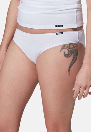 Primer plano de una persona que lleva ropa interior blanca y una camiseta blanca, mostrando un tatuaje tribal negro en la parte superior del muslo.