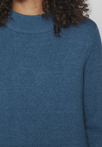 Person med lockigt mörkt hår iförd en blå stickad tröja med ribbat rundhals, visad från halsen till mitten av överkroppen.