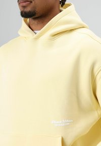 Geel hoodiesweater gemaakt van zachte stof. Heeft een zak aan de voorkant en geborduurde tekst op de linkerzoom. Soepele textuur en een comfortabele pasvorm.