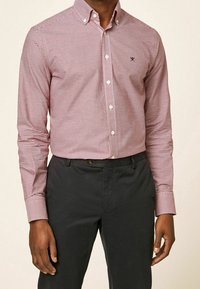 Czerwona i biała koszula w kratę na guziki z długimi rękawami, w połączeniu z ciemnymi spodniami. Na piersi znajduje się małe logo.