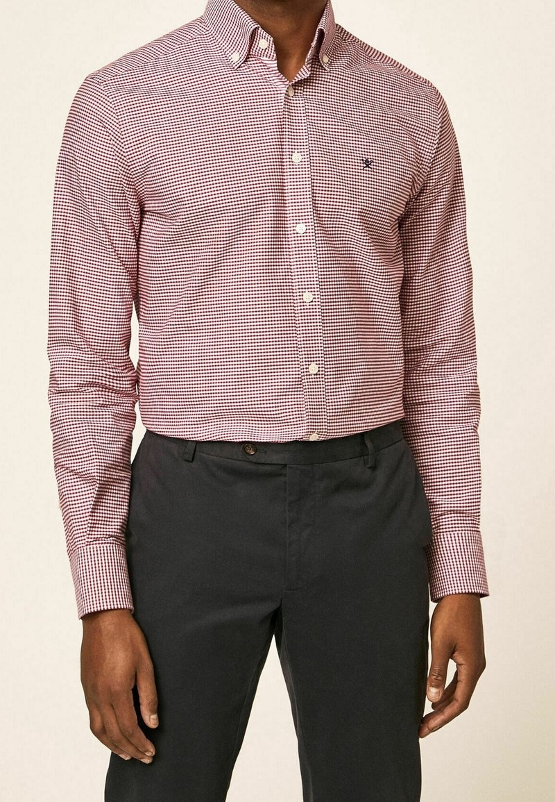 Czerwona i biała koszula w kratę na guziki z długimi rękawami, w połączeniu z ciemnymi spodniami. Na piersi znajduje się małe logo.
