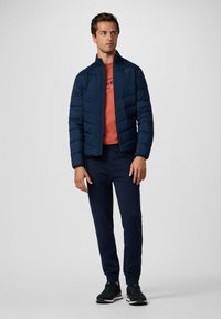 Marineblauwe pufferjack met een hoge kraag en een doorgestikt ontwerp, gecombineerd met een oranje shirt en donkerblauwe joggers. Zwarte sneakers maken de look compleet.