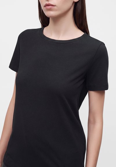 Femme portant un t-shirt noir uni à manches courtes avec un col rond sur un fond clair uni.