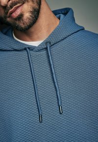 Felpa blu con texture e motivo a diamante, con lacci regolabili e punte in argento. Sotto si intravede una maglietta bianca a girocollo.