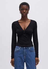 Zwarte lange mouwen top met een V-hals en een twistdetail op de borst. Strakke pasvorm met geribbelde textuur, gecombineerd met hoge, blauwe jeans.